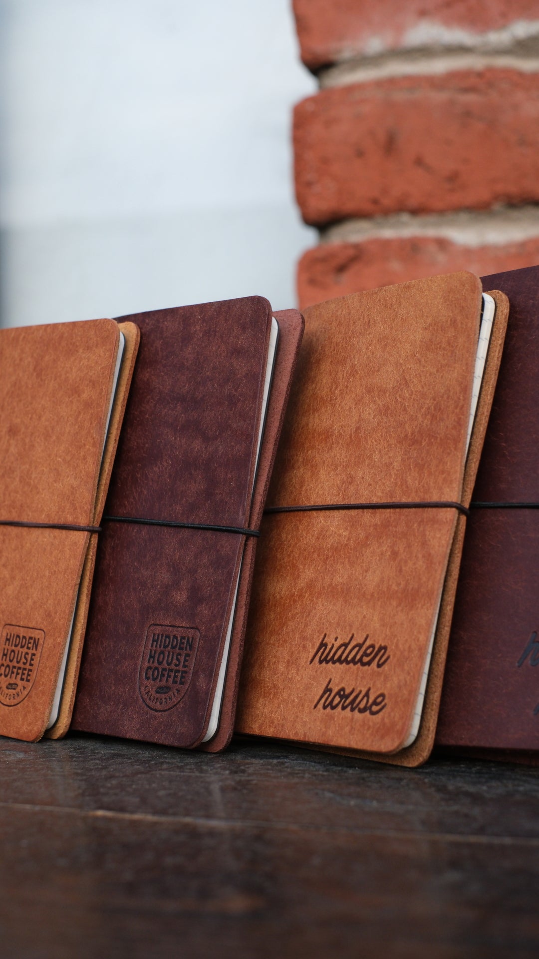 Picture of four leather journals stacked, with variation of colors and two different Hidden House Coffee logos.
