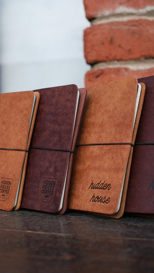 Picture of four leather journals stacked, with variation of colors and two different Hidden House Coffee logos.