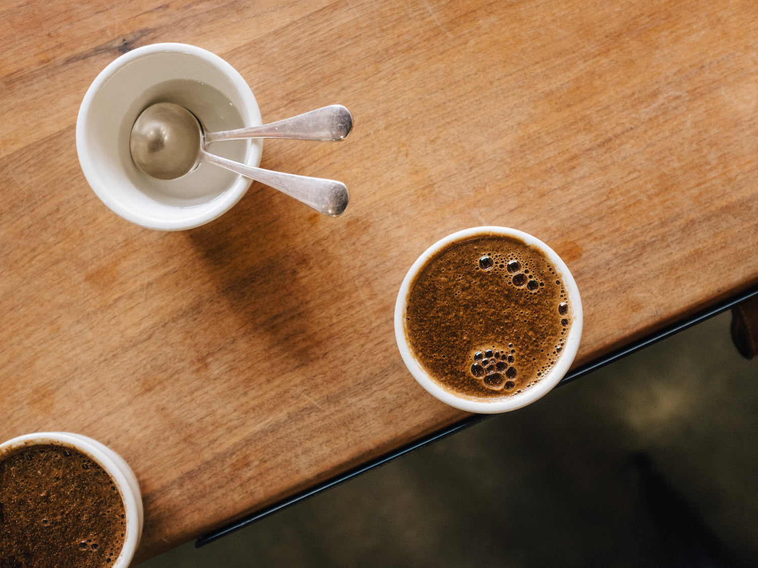  Image of coffee inside of cupping cups on top of a table. There are also cupping spoons in a cup filled with clean water.