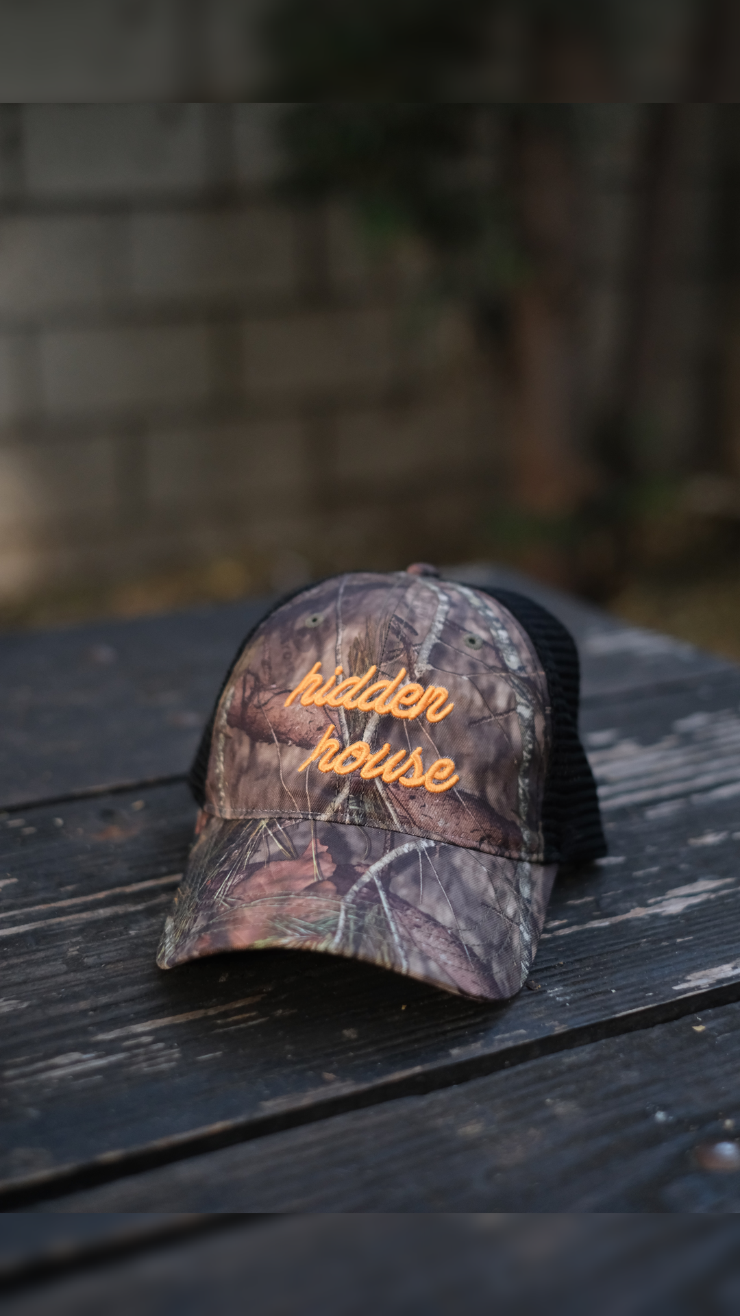 Picture of camo trucker hat with hidden house logo embroidered in yellow.