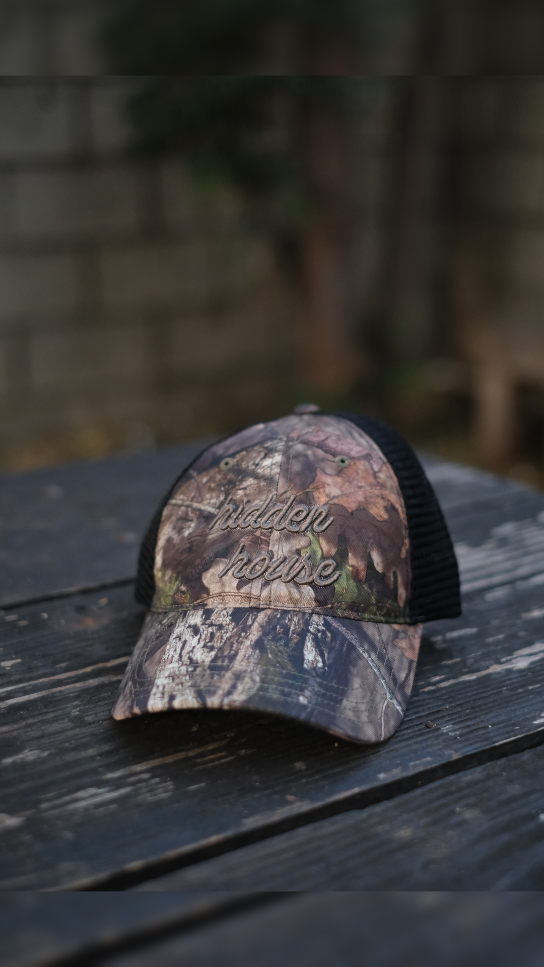 Picture of camo trucker hat with hidden house logo embroidered in gray.