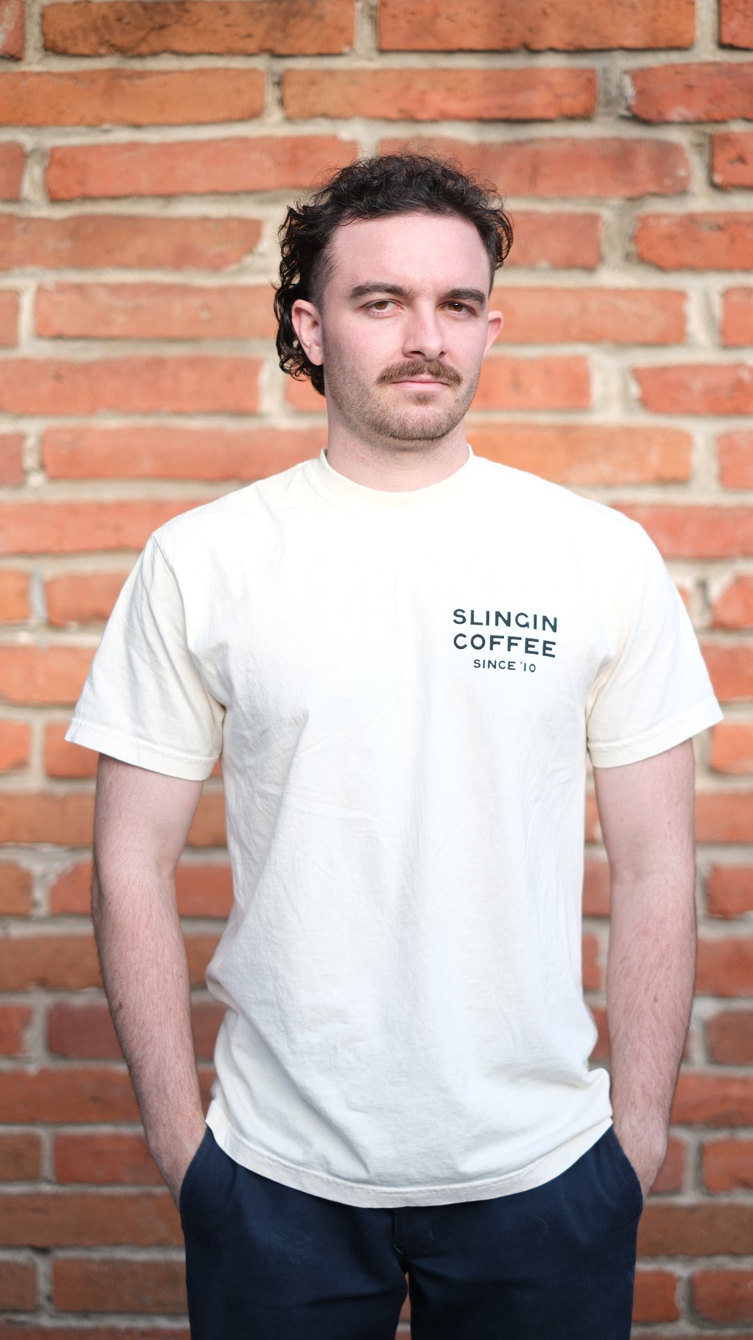 Photo of model wearing el ray tee. On front of shirt, it is printed with "slingin coffee since '10"