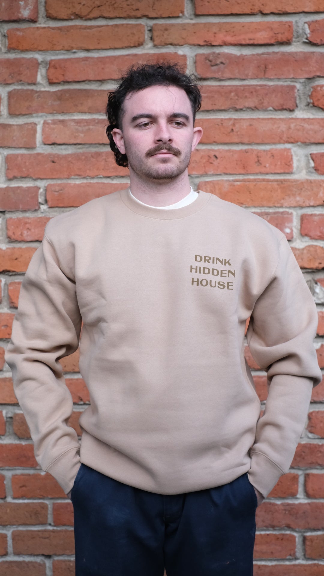 Model wearing hidden house crew neck sweatshirt that is printed with "drink hidden house" on the front.