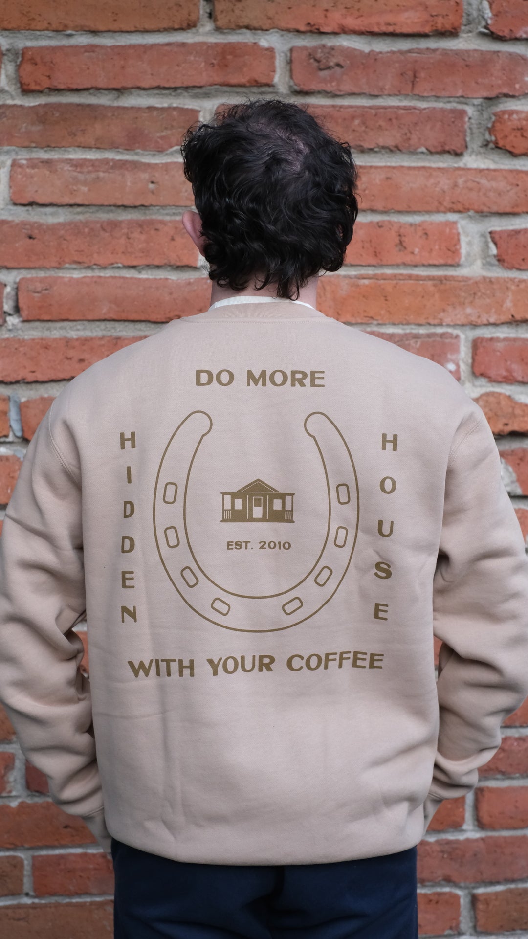 Model showing Back of crew neck sweatshirt featuring HHC artwork that says "Do more with your coffee, hidden house, established 2010".
