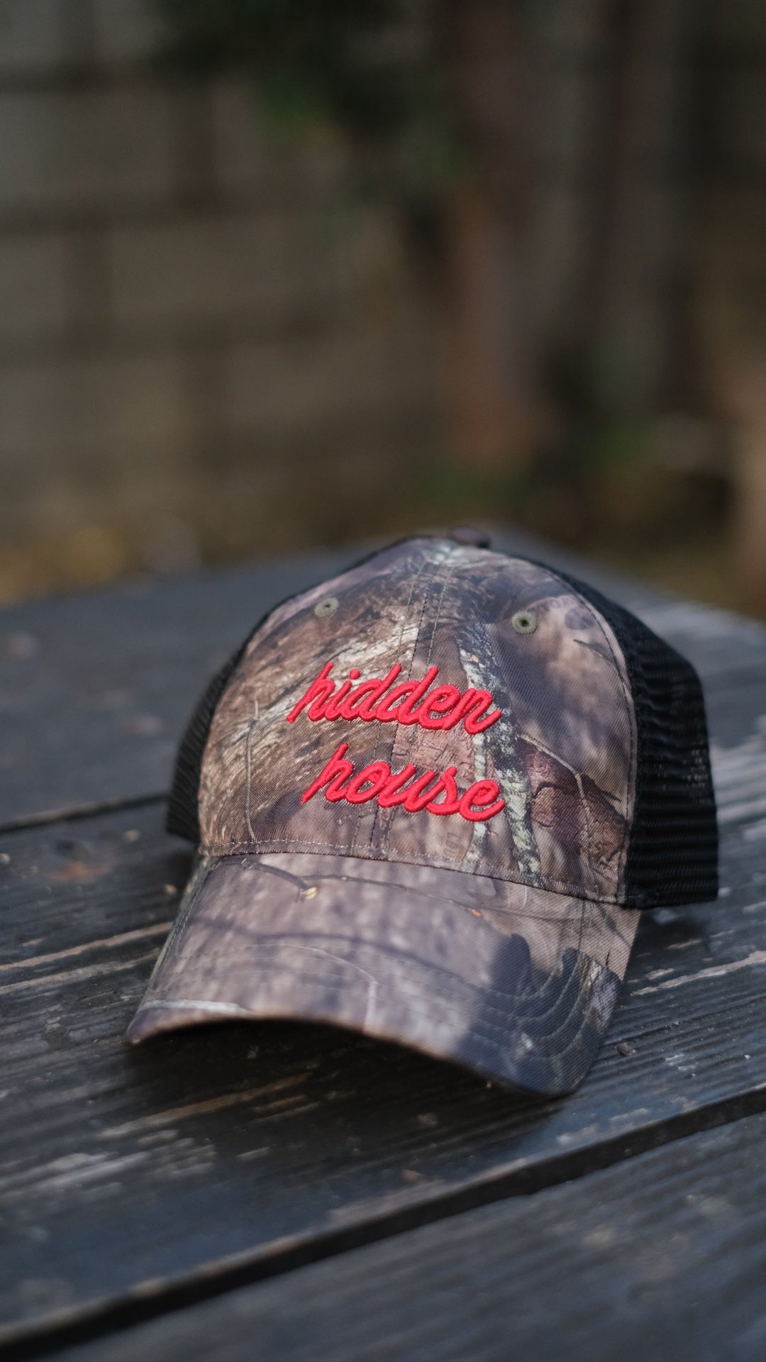 Picture of camo trucker hat with hidden house logo embroidered in red.